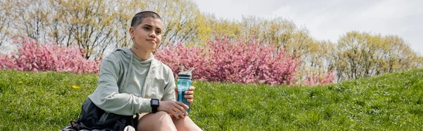 Caminhante feminina de cabelos curtos e tatuado segurando garrafa de esportes enquanto sentado perto da mochila na colina gramada com paisagem cênica no fundo, percorrendo paisagem cênica, banner — Fotografia de Stock