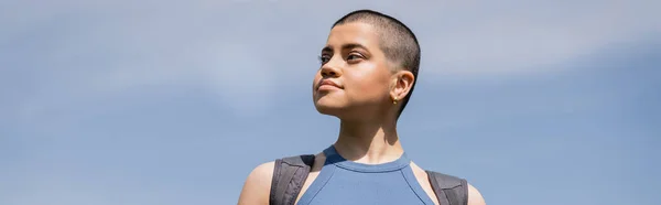 Joven turista femenina segura y de pelo corto con mochila mirando hacia otro lado mientras está de pie con el cielo azul en el fondo, concepto de viaje de senderismo en solitario, pancarta, verano - foto de stock