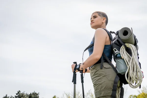 Low angle view of young short haired female backpacker with fitness tracker and backpack looking away while holding trekking poles with sky at background, solo hiking journey concept — Stock Photo