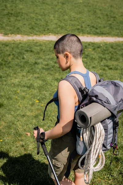 Vue latérale d'une jeune randonneuse aux cheveux courts et tatouée avec sac à dos et tracker de fitness tenant un bâton de trekking tout en marchant sur une pelouse herbeuse en arrière-plan, concept de voyage de randonnée en solo — Photo de stock