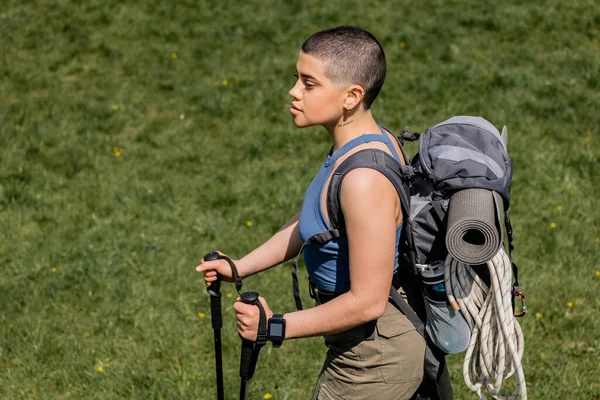 Joven excursionista femenina de pelo corto y tatuada con mochila y rastreador de fitness que sostiene bastones de trekking y camina sobre césped cubierto de hierba en el fondo, concepto de viaje de senderismo en solitario, verano - foto de stock