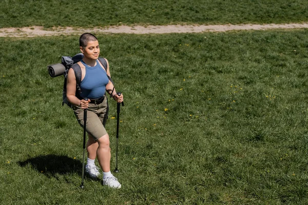 High angle view of smiling young short haired female hiker with backpack holding trekking poles and walking on grassy lawn at background, solo hiking journey concept, summer — Stock Photo