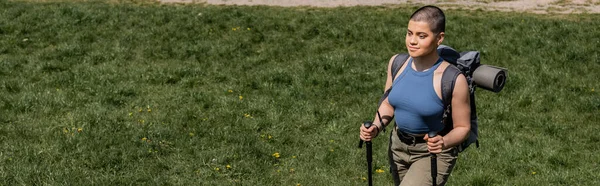 Vue grand angle du jeune routard femelle aux cheveux courts avec sac à dos souriant et tenant des bâtons de trekking tout en marchant sur un pré herbeux en arrière-plan, concept de voyage de randonnée en solo, bannière — Photo de stock