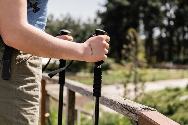 Vista recortada de una joven mujer tatuada que sostiene bastones de trekking mientras está de pie cerca de una valla de madera con la naturaleza al fondo, reconectándose con usted mismo en la naturaleza, Traducción del tatuaje: amor - foto de stock