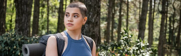 Young short haired female traveler with backpack and fitness mat looking away while standing in blurred green forest, reconnecting with yourself in nature concept, banner — Stock Photo