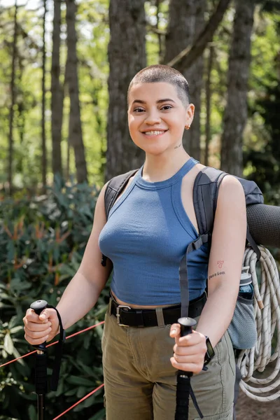 Portrait of smiling young short hired woman hiker with backpack holding trekking poles and looking at camera while standing in blurred green forest, reconnecting with yourself in nature concept — Stock Photo