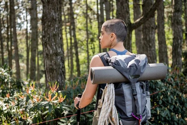 Вид сбоку на молодую коротковолосую и татуированную женщину-туристку с рюкзаком, держащую шесты и смотрящую в сторону, стоя в зеленом лесу, воссоединяющуюся с собой в концепции природы — стоковое фото