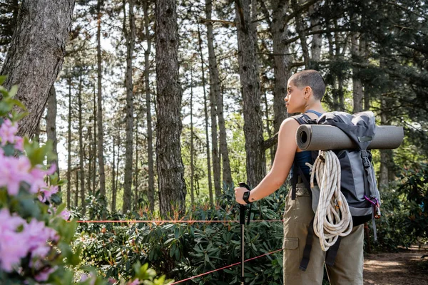 Вид сбоку молодой коротковолосой туристки с рюкзаком и туристическим снаряжением, держащей походный столб и стоящей в зеленом лесу, воссоединяющейся с собой в природе, лето — стоковое фото