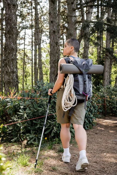 Vista lateral de una joven viajera de pelo corto con mochila sosteniendo poste de trekking y senderismo en el verde bosque de verano, reconectando con usted mismo en concepto de naturaleza - foto de stock
