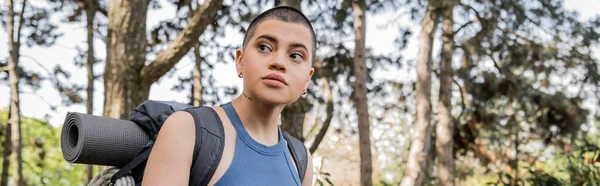 Portrait of young short haired and tattooed female tourist with backpack looking away while standing in blurred green summer forest at background, hiking for health and wellness concept, banner — Stock Photo