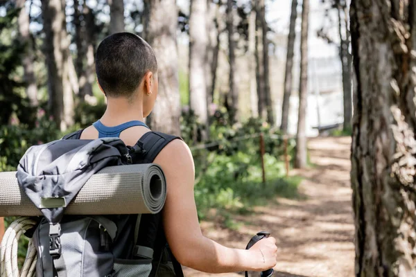 Vista posterior de una joven turista de pelo corto y tatuada con mochila y colchoneta de fitness sosteniendo el poste de trekking mientras camina en un bosque verde borroso, Traducción del tatuaje: amor - foto de stock