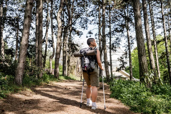Вид сбоку на молодую коротковолосую женщину с рюкзаком и туристическим снаряжением, держащую в руках походные шесты, стоя в размытом зеленом лесу, путешествуя ради здоровья и хорошего самочувствия — стоковое фото
