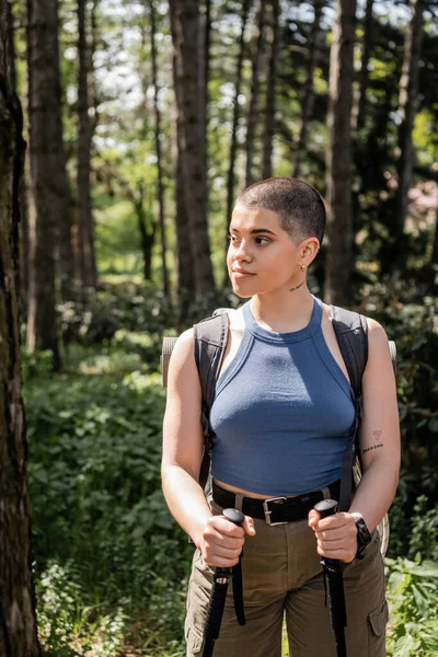 Молода і впевнена короткошерста татуйована жінка з рюкзаком, що тримає пішохідні полюси і дивиться, стоячи в літньому лісі, пішохідна концепція здоров'я та оздоровлення — стокове фото