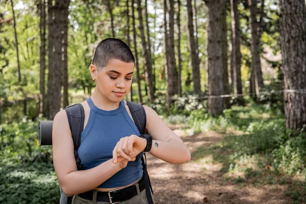 Young short haired and tattooed woman tourist with backpack looking at fitness tracker while hiking in blurred summer forest at background, wanderlust vibes concept — Stock Photo