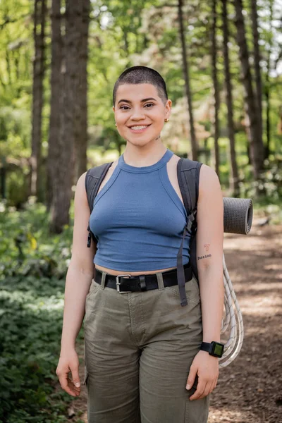 Fröhliche, kurzhaarige und tätowierte Wanderin mit Rucksack und Fitness-Tracker blickt in die Kamera, während sie im Hintergrund im grünen Sommerwald steht, Fernweh kommt auf. — Stockfoto