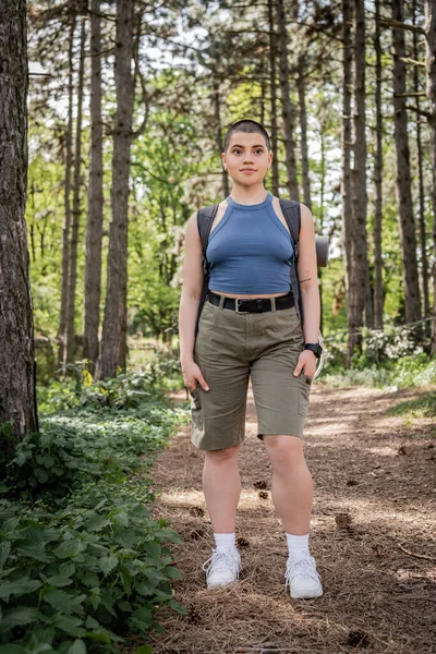Junge und selbstbewusste Wanderin mit Rucksack und Smartwatch, die wegschaut und die Hände in Shorts hält, während sie im grünen Sommerwald steht, weckt Fernweh. — Stockfoto