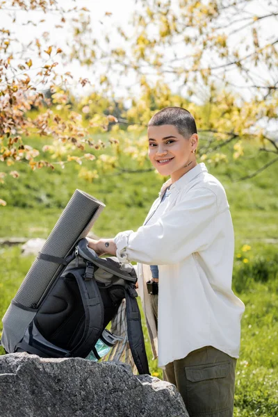 Smiling short haired and tattooed woman hiker in casual clothes looking at camera near backpack on stone with blurred scenic landscape at background , confident female explorer, summer — Stock Photo