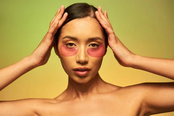 Hautpflege-Kampagne, junge asiatische Frau mit brünetten Haaren und sauberer Haut posiert mit Händen in Kopfnähe auf grünem Hintergrund, nackten Schultern, feuchtigkeitsspendenden Augenflecken, glühender Haut — Stockfoto