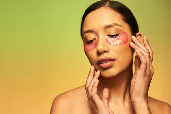 Campanha de cuidados com a pele, sensual mulher asiática com cabelo morena e pele limpa posando com as mãos perto do rosto no fundo verde, ombros nus, manchas hidratantes nos olhos, pele brilhante — Fotografia de Stock