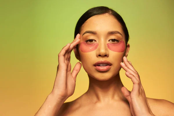 Schönheitsprodukt, junge asiatische Frau mit brünetten Haaren und sauberer Haut posiert und schaut in die Kamera auf grünem Hintergrund, nackte Schultern, feuchtigkeitsspendende Augenflecken, glühende Haut — Stockfoto