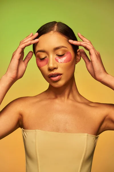 Модель красоты, светящаяся кожа, молодая азиатская женщина с брюнетками и чистой кожей позирует и смотрит вниз на зеленый фон, голые плечи, увлажняющие повязки на глаза, кожа продукт — стоковое фото