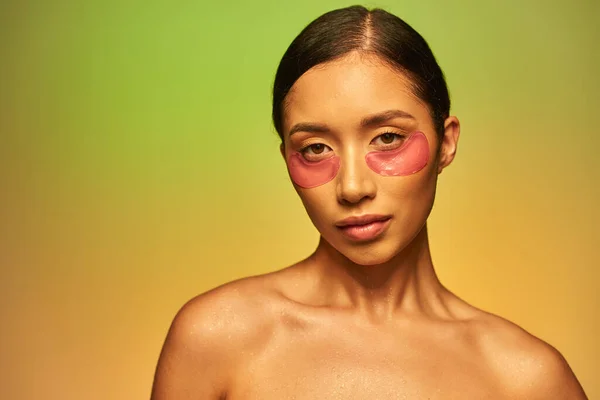 Campanha skincare, jovem mulher asiática com cabelo morena e ombros nus posando e olhando para a câmera em fundo verde, cuidados com o rosto, manchas hidratantes nos olhos, pele brilhante — Fotografia de Stock