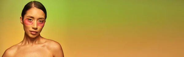 Campagna di cura della pelle, giovane donna asiatica con capelli castani e spalle nude in posa e guardando la fotocamera su sfondo verde, cura del viso, macchie idratanti sugli occhi, pelle luminosa, banner — Foto stock