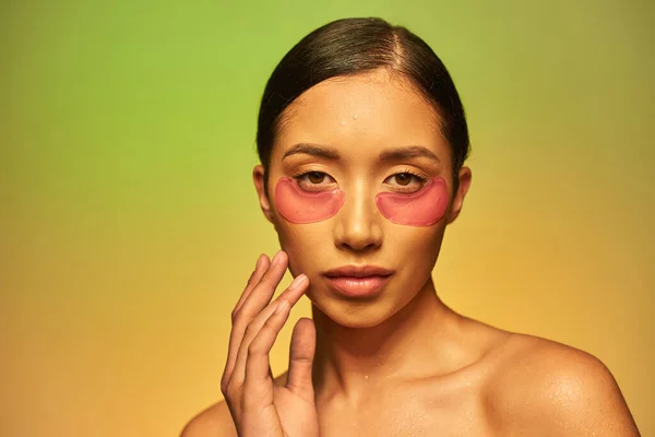Campanha de cuidados com a pele, jovem mulher asiática com cabelo morena e pele limpa tocando bochecha e olhando para a câmera no fundo verde, ombros nus, manchas hidratantes nos olhos, pele brilhante — Fotografia de Stock