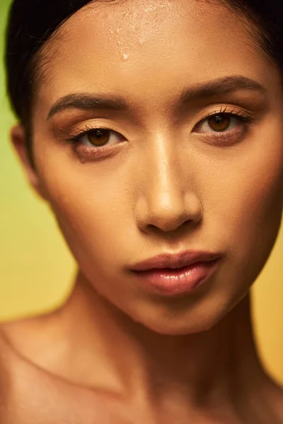 Gotas de agua en la cara, primer plano de la joven mujer asiática con la piel húmeda mirando a la cámara en el fondo verde, hidratación de la piel, campaña de belleza, perfección, bienestar, conceptual - foto de stock