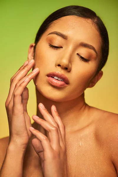 Gotas de agua en la cara, primer plano de mujer asiática joven con la piel húmeda tocando la cara sobre fondo verde, ojos cerrados, hidratación de la piel, campaña de belleza, perfección, bienestar, conceptual - foto de stock
