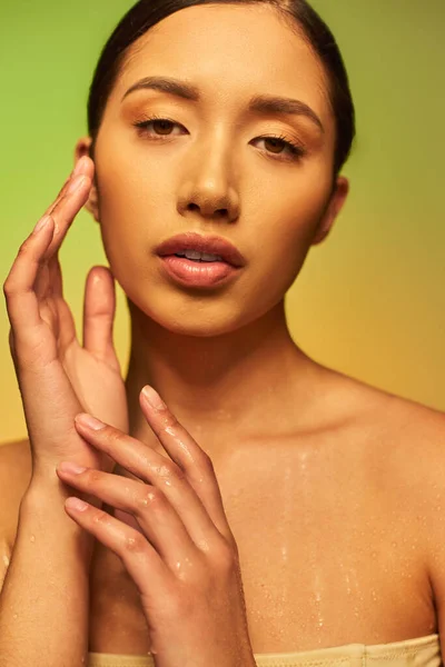 Hydration, young asian woman with bare shoulders and wet body posing on gradient background, touching face, looking at camera, skincare campaign, beauty model, brunette hair, glowing skin — Stock Photo