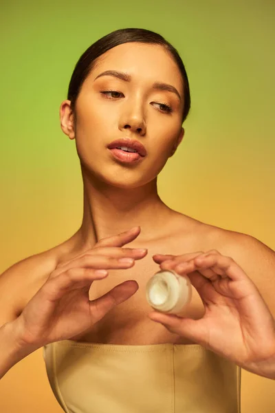 Campanha de beleza, jovem mulher asiática com ombros nus segurando frasco cosmético com creme facial no fundo verde, cabelo morena, indústria de beleza, pele brilhante, conceito de cuidados com a pele — Fotografia de Stock