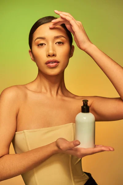 Presentación del producto, mujer asiática joven con hombros desnudos sosteniendo botella cosmética con producto de belleza y mirando a la cámara sobre fondo verde, cabello moreno, piel brillante - foto de stock