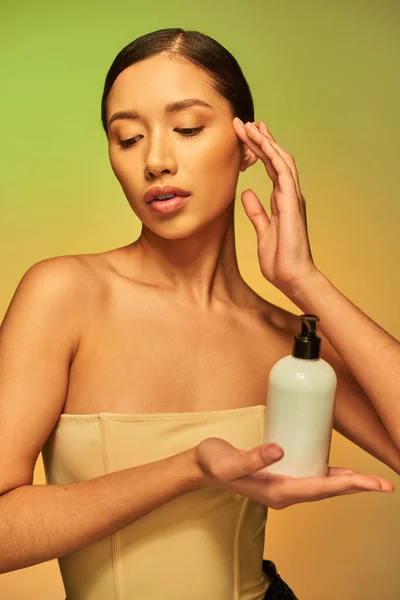 Presentazione del prodotto, prodotto di bellezza, giovane donna asiatica con spalle nude che tiene la bottiglia cosmetica con lozione per il corpo e posa su sfondo verde, capelli castani, pelle luminosa — Foto stock