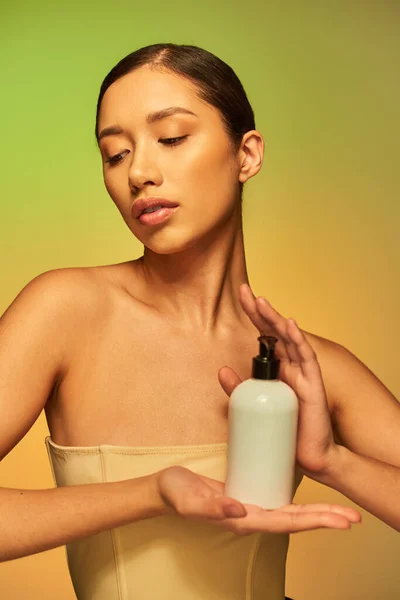 Présentation du produit, soin de la peau produit, jeune femme asiatique aux épaules nues tenant bouteille cosmétique avec lotion corporelle et posant sur fond vert, peau éclatante, cheveux bruns — Photo de stock