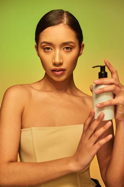 Présentation du produit, soin de la peau produit, jeune femme asiatique aux épaules nues tenant bouteille cosmétique avec lotion corporelle et posant sur fond vert, concept de peau éclatante — Photo de stock