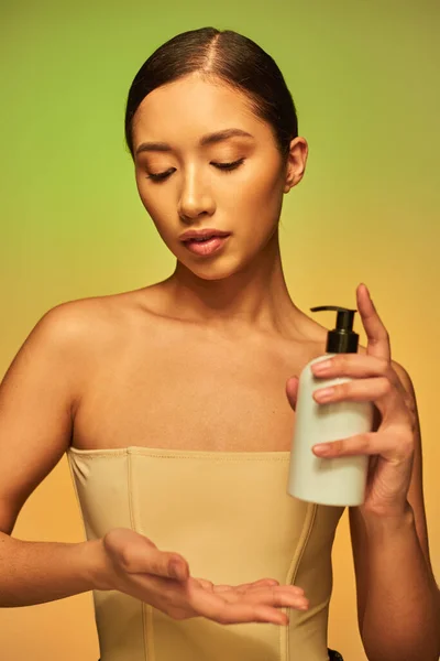 Presentación del producto, producto de cuidado de la piel, mujer asiática joven con hombros desnudos sosteniendo botella cosmética y posando sobre fondo verde, piel brillante, cabello moreno - foto de stock