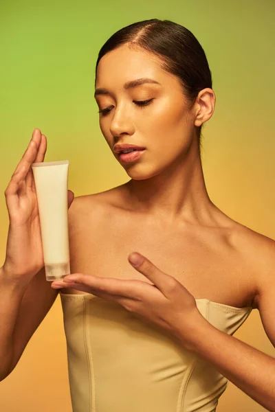Cura della pelle, presentazione del prodotto, pelle giovane, giovane donna asiatica con spalle nude che tiene il tubo cosmetico e posa su sfondo verde, pelle luminosa, capelli castani — Foto stock