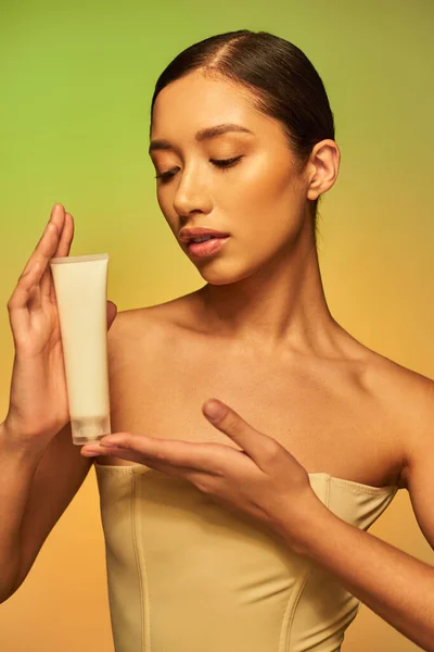 Soins de la peau, présentation de produits, peau jeune, jeune femme asiatique aux épaules nues tenant le tube cosmétique et posant sur fond vert, peau éclatante, cheveux bruns — Photo de stock