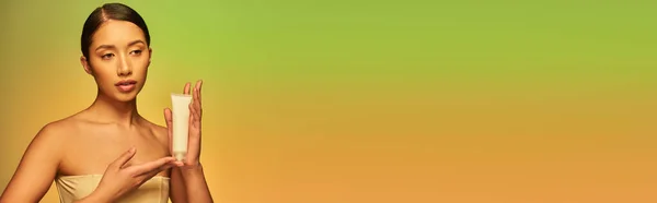 Cuidado de la piel, presentación del producto, mujer asiática joven con hombros desnudos sosteniendo tubo cosmético y posando sobre fondo verde, piel brillante, cabello moreno, piel juvenil, bandera - foto de stock