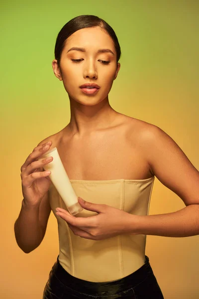 Cuidados com a pele, apresentação do produto, jovem mulher asiática com ombros nus segurando tubo cosmético com creme e posando em fundo verde, pele brilhante, cabelo morena, pele jovem — Fotografia de Stock