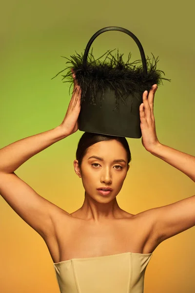 Belleza y estilo, morena mujer asiática con hombros desnudos posando con monedero de plumas en la cabeza sobre fondo verde, degradado, declaración de moda, piel brillante, belleza natural, modelo joven - foto de stock