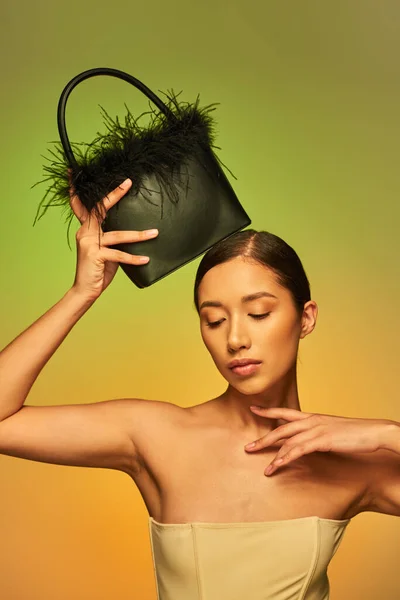 Schönheit und Stil, brünette asiatische Frau mit nackten Schultern posiert mit Federhandtasche auf grünem Hintergrund, Hand nahe Gesicht, Steigung, Mode-Statement, glühende Haut, natürliche Schönheit, junges Modell — Stockfoto