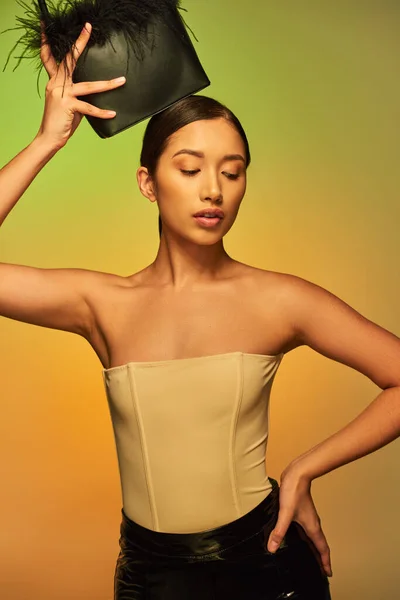 Beauté et de style, femme asiatique brune aux épaules nues posant avec sac à main en plumes et la main sur la hanche sur fond vert, dégradé, déclaration de mode, peau éclatante, beauté naturelle, jeune modèle — Photo de stock