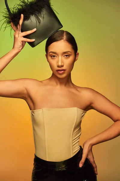 Modewahl, brünette asiatische Frau mit nackten Schultern posiert mit Federtasche und Hand auf Hüfte auf grünem Hintergrund, Steigung, modisches Statement, glühende Haut, natürliche Schönheit, junges Model — Stockfoto