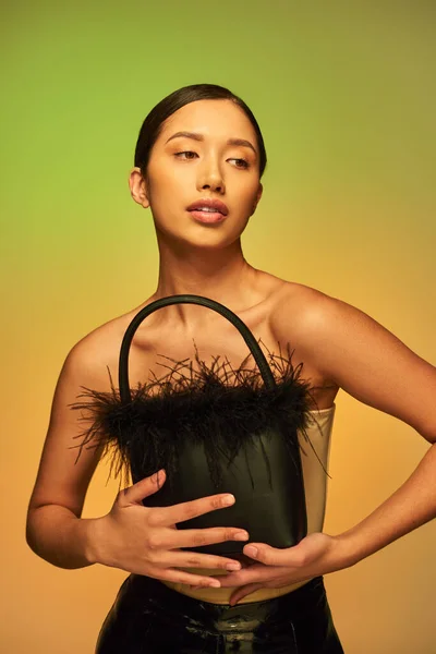 Modewahl, brünette asiatische Frau mit nackten Schultern posiert mit Federtasche auf grünem Hintergrund, Steigung, Mode nach vorne, glühende Haut, natürliche Schönheit, junges Model — Stockfoto