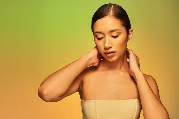Fotografia de beleza, mulher asiática com cabelo morena e ombros nus posando no fundo gradiente, verde e laranja, cuidados com a pele, pele brilhante, beleza natural, modelo jovem — Fotografia de Stock