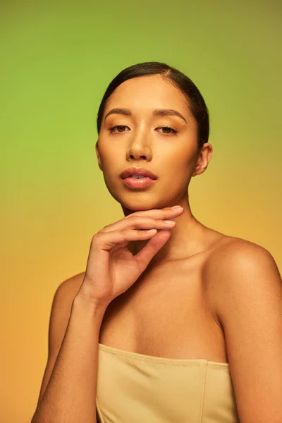 Краса і догляд за шкірою, азіатська жінка з брюнеткою волоссям і голими плечима позує на градієнтному фоні, зелений і оранжевий, догляд за шкірою, сяюча шкіра, природна краса, молода модель — стокове фото