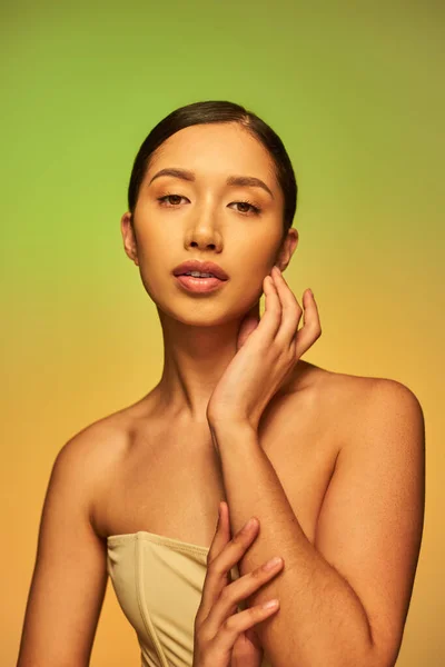 Schönheitsfotografie, hübsche asiatische Frau mit brünetten Haaren und nackten Schultern posiert auf Gradienten Hintergrund, grün und orange, Hautpflege, glühende Haut, natürliche Schönheit, junges Modell — Stockfoto