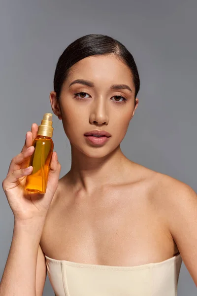 Product presentation, skin care, young asian model with brunette hair holding cosmetic bottle with oil on grey background, glowing and heathy skin, beauty campaign, facial treatment concept — Stock Photo